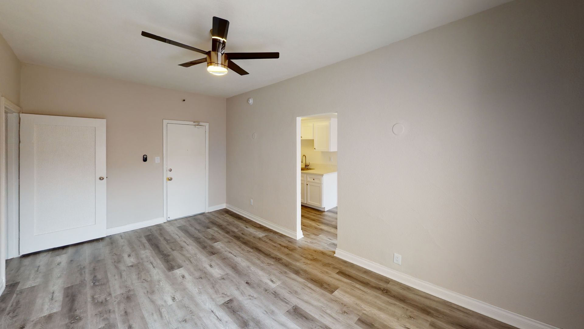 Empty living room with light grey walls, dark wood-look flooring, and an open doorway to a kitchen.