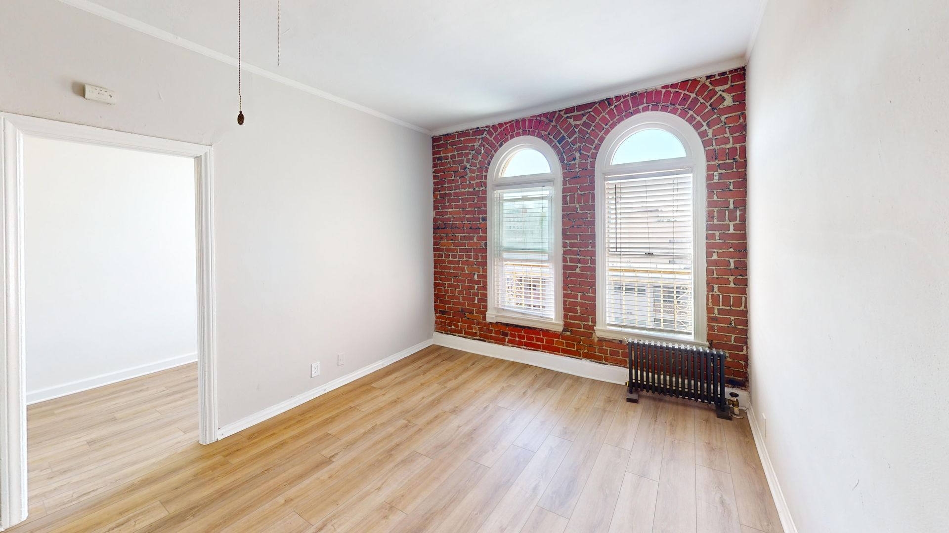 Empty room with exposed brick wall, arched windows, and light wood flooring.