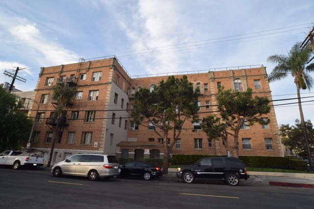Four-story brick apartment building on a city street, cars parked out front, trees, and blue sky.