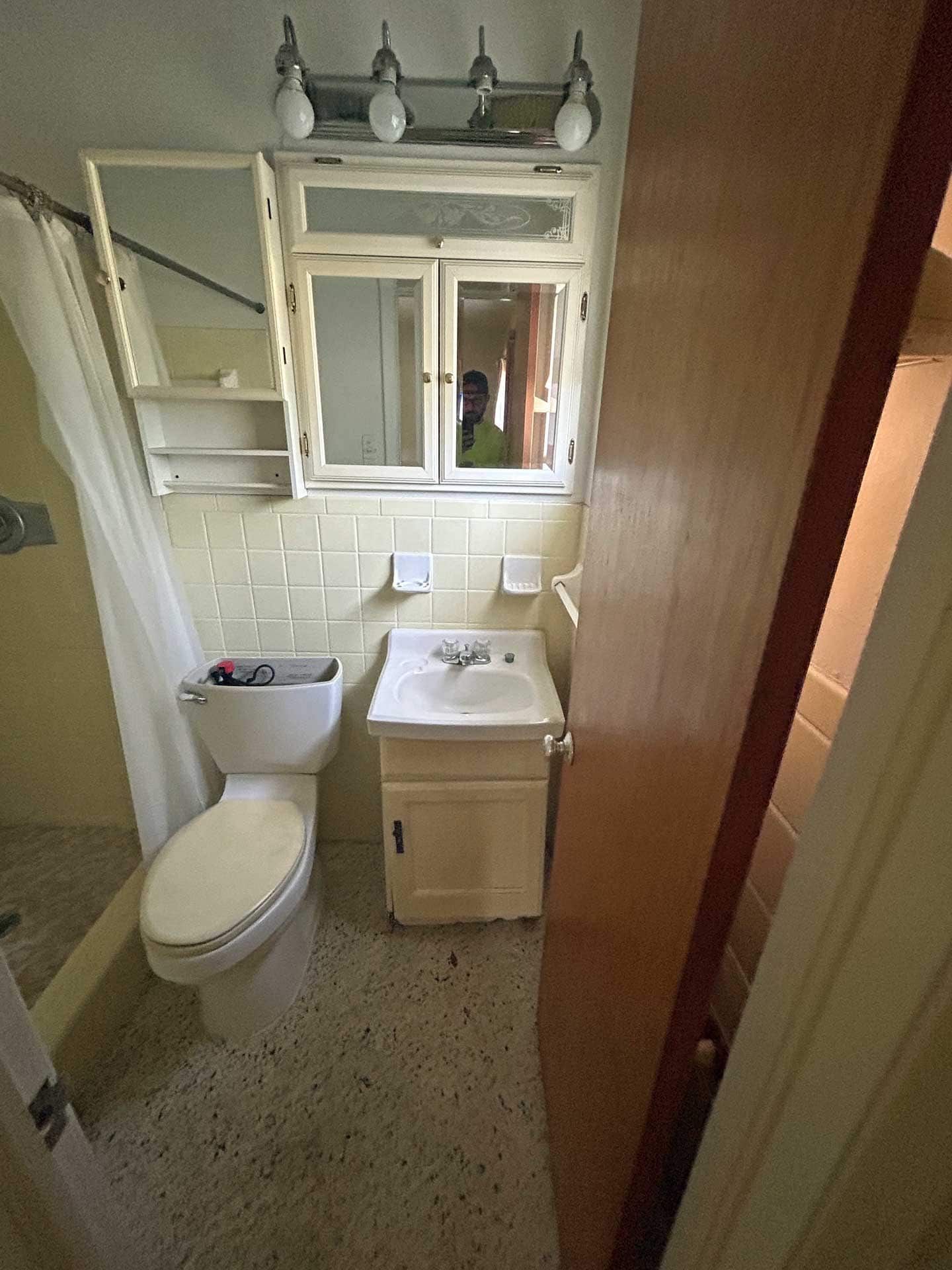 Small bathroom with a toilet, sink, medicine cabinet, and shower. Beige tiles, light fixtures, and open door.