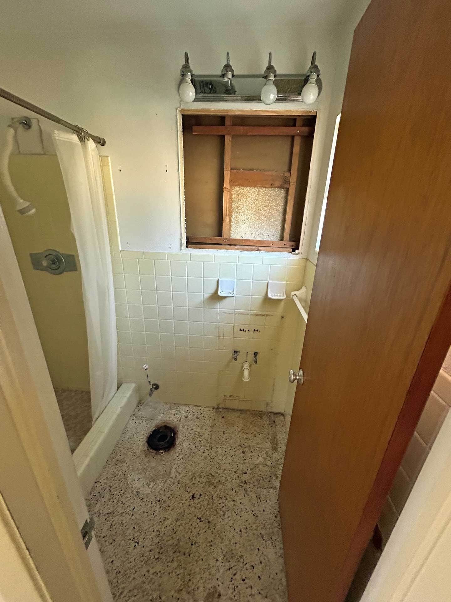 Bathroom interior with visible wall damage, old tile, and exposed plumbing. A shower and door are also visible.