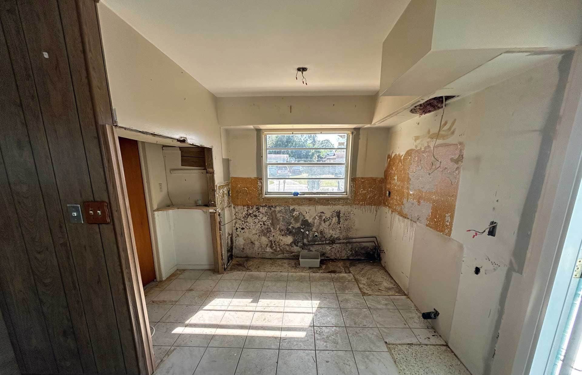 Interior view of a gutted kitchen with visible damage, window, and open doorway.