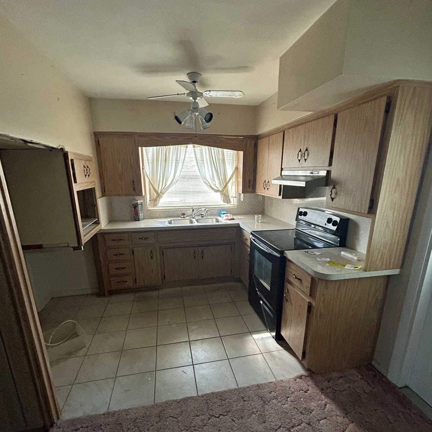 U-shaped kitchen with wood cabinets, outdated appliances, a window with curtains, and tile flooring.