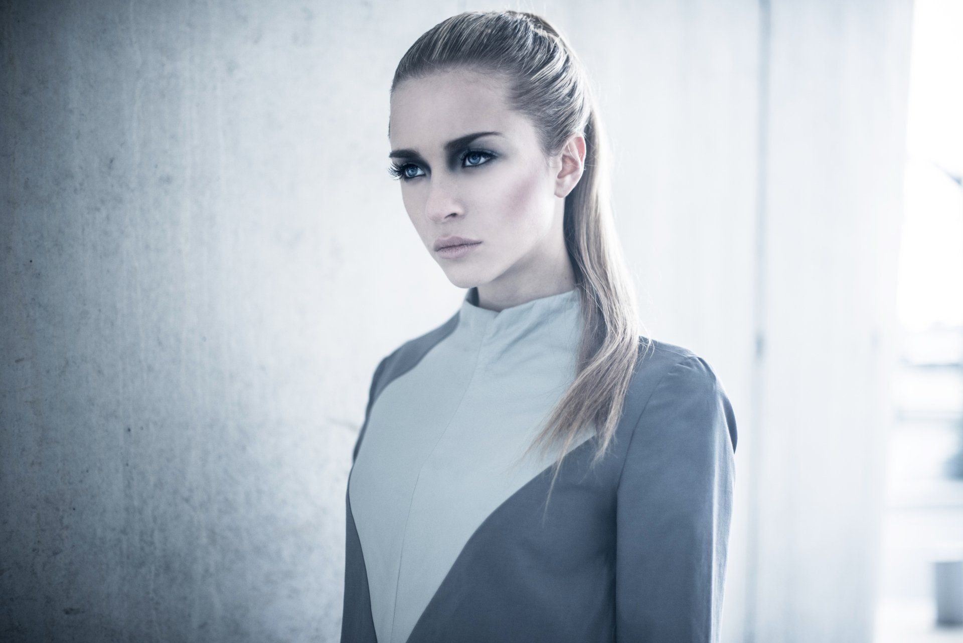 Eine Frau in einem grau-weißen Kleid steht vor einer Betonmauer.