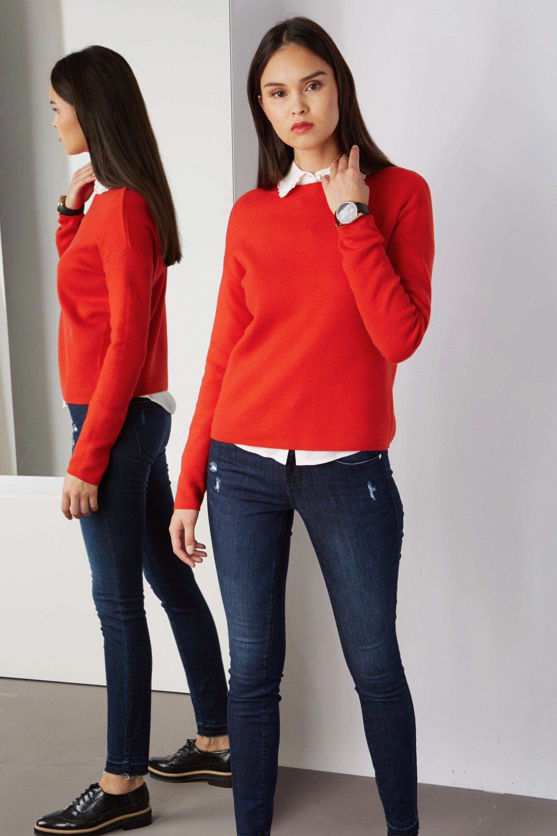 Eine Frau in einem roten Pullover und blauen Jeans steht vor einem Spiegel.