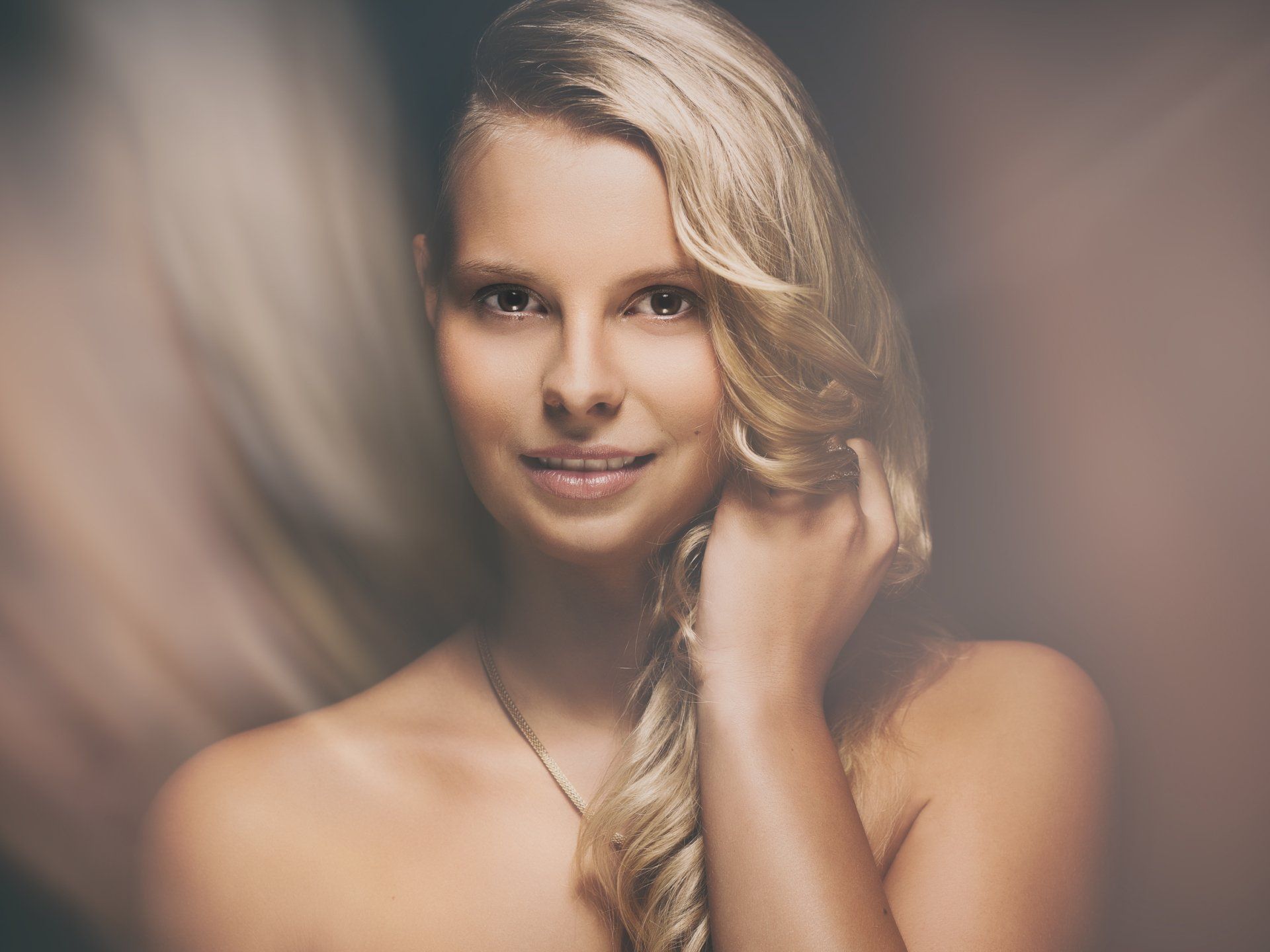 Blonde Frau mit schulterlangem Haar, lächelt, Hand in der Nähe des Kopfes und Halskette; weicher Hintergrund.