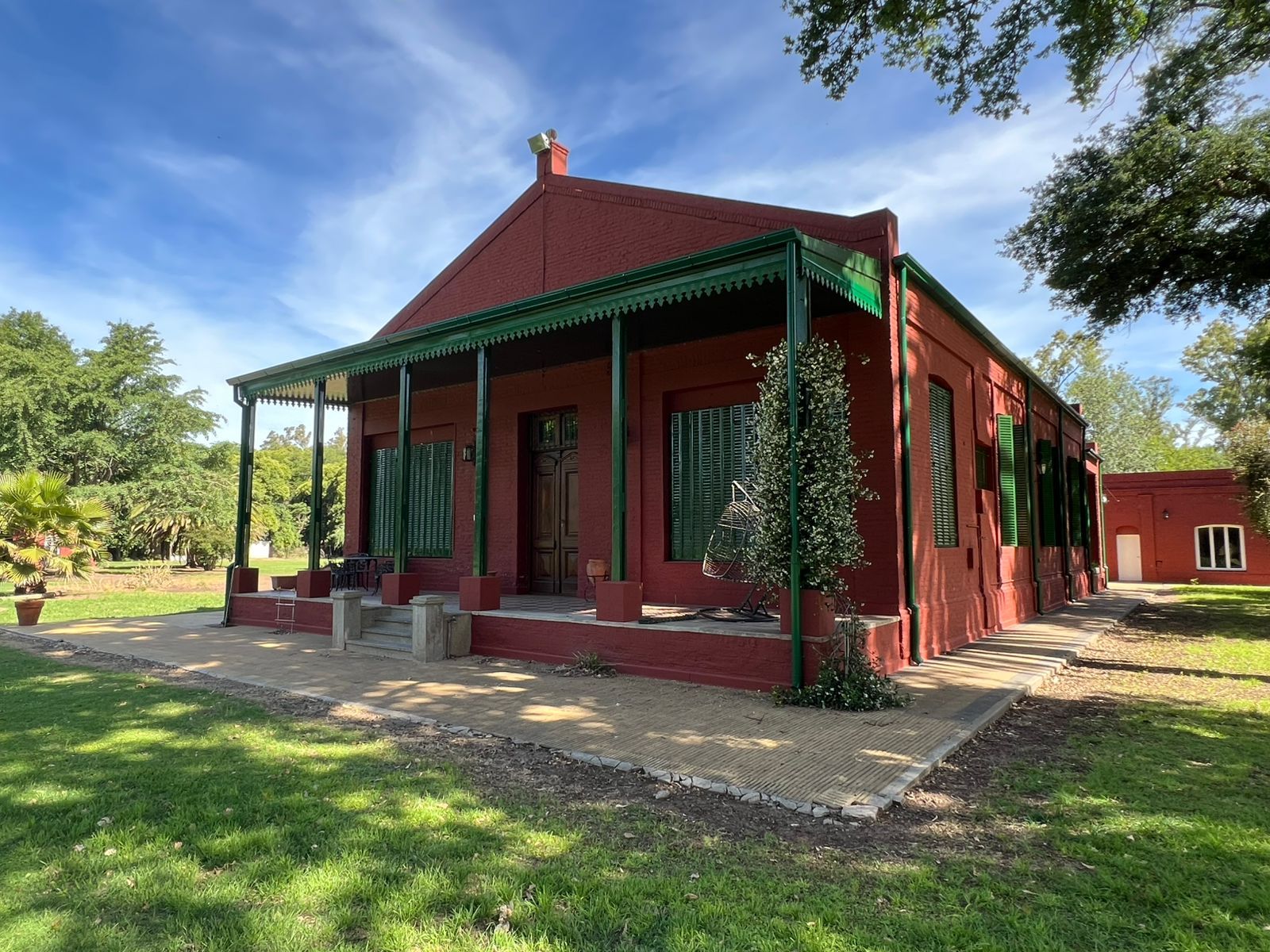 Casita de madera roja con porche revestido de verde, rodeada de árboles y césped bajo un cielo azul.