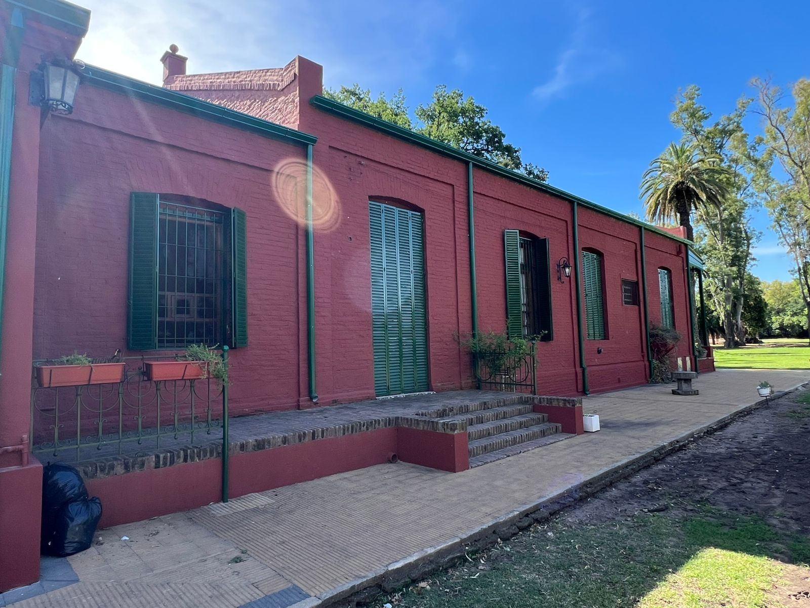 Edificio de ladrillo rojo con contraventanas y puertas verdes a lo largo de un sendero soleado con árboles cerca.