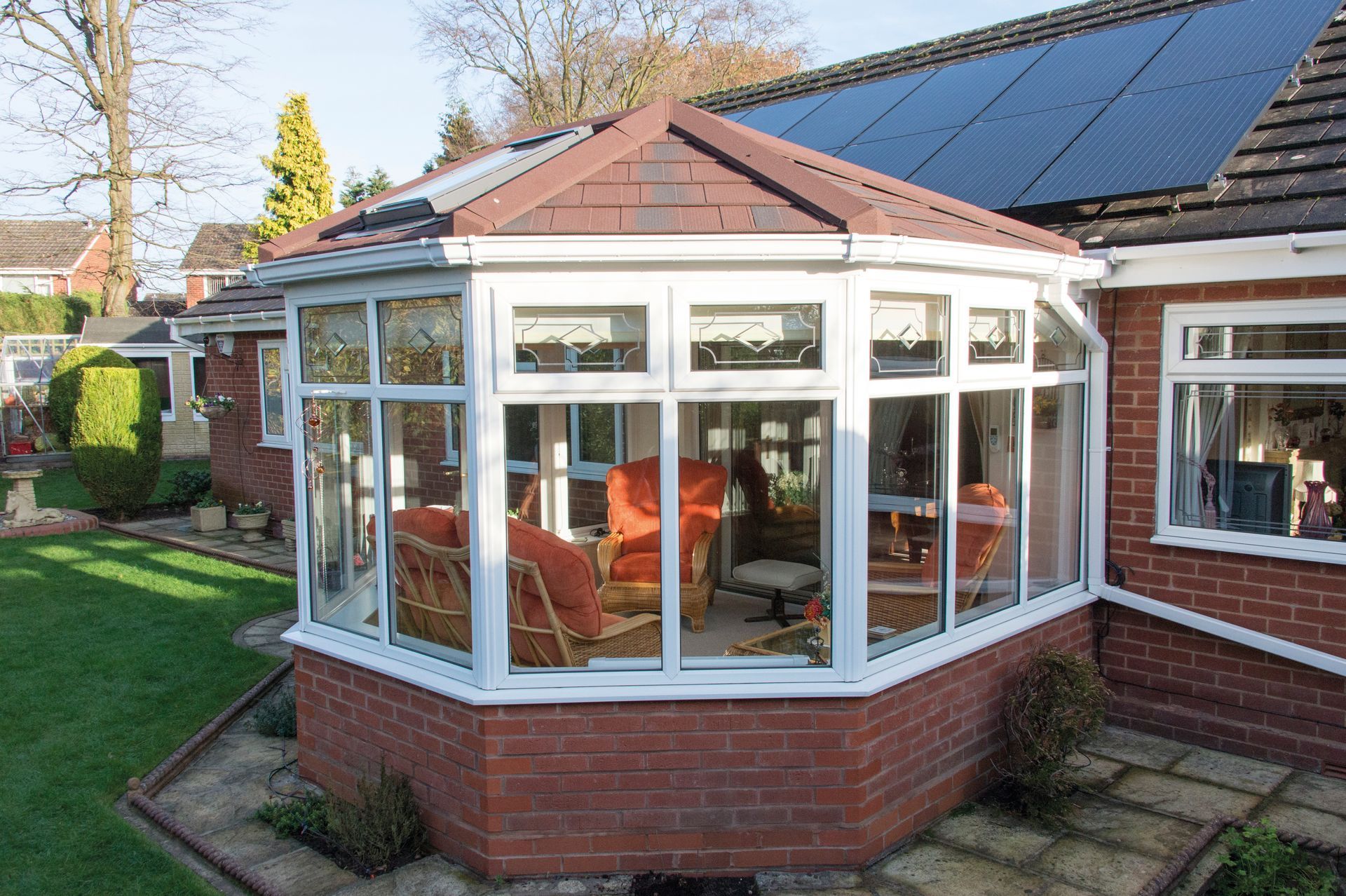 Conservatory with brick base, glass windows, tiled roof, and furnished interior, attached to a red brick house.