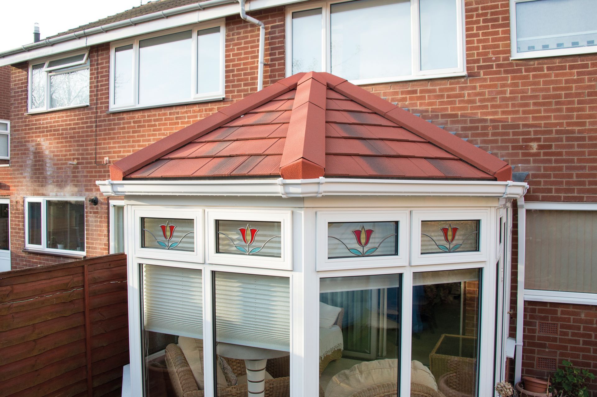 Conservatory with red tile roof, white frames, and stained-glass accents, attached to a brick house.