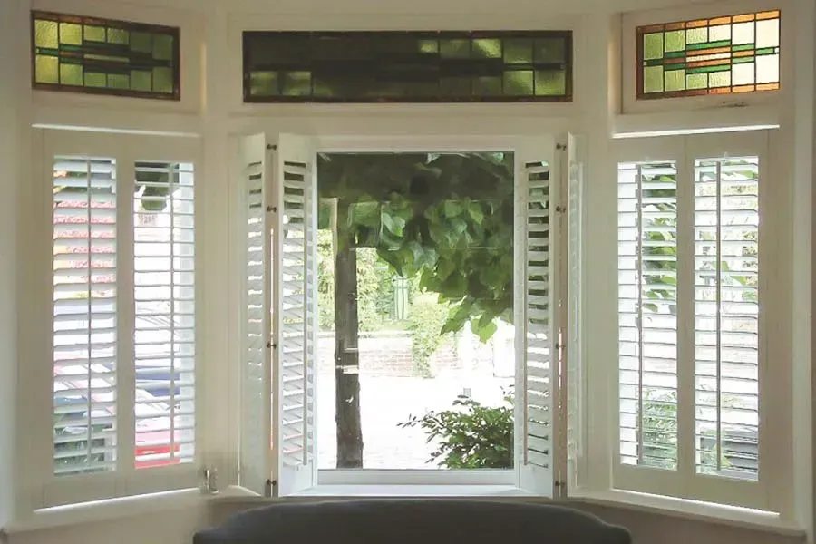Bay window with white shutters, stained glass transom, and view of greenery.