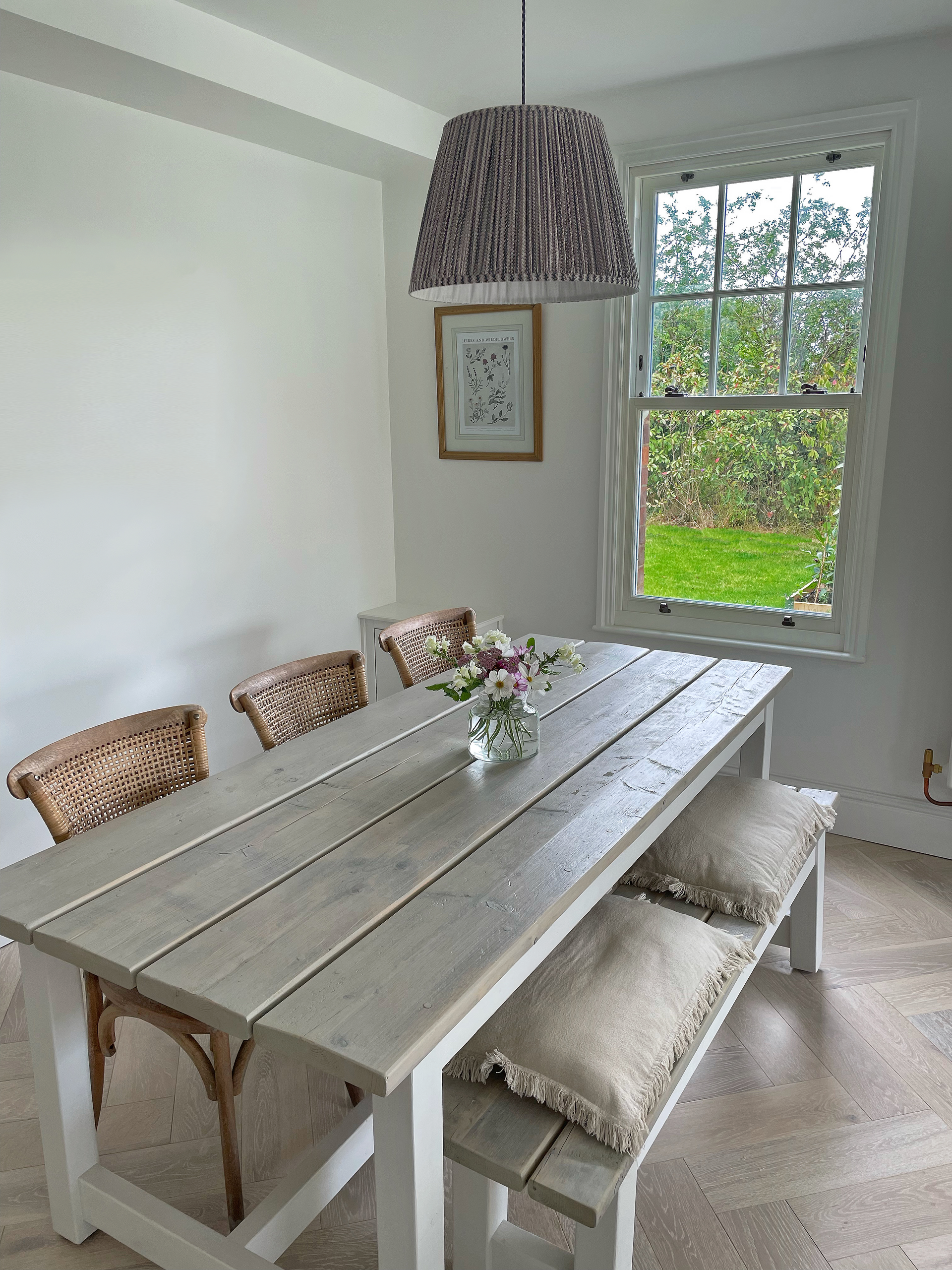 Dining room with a long gray table, benches, and chairs under a woven lampshade by a window.