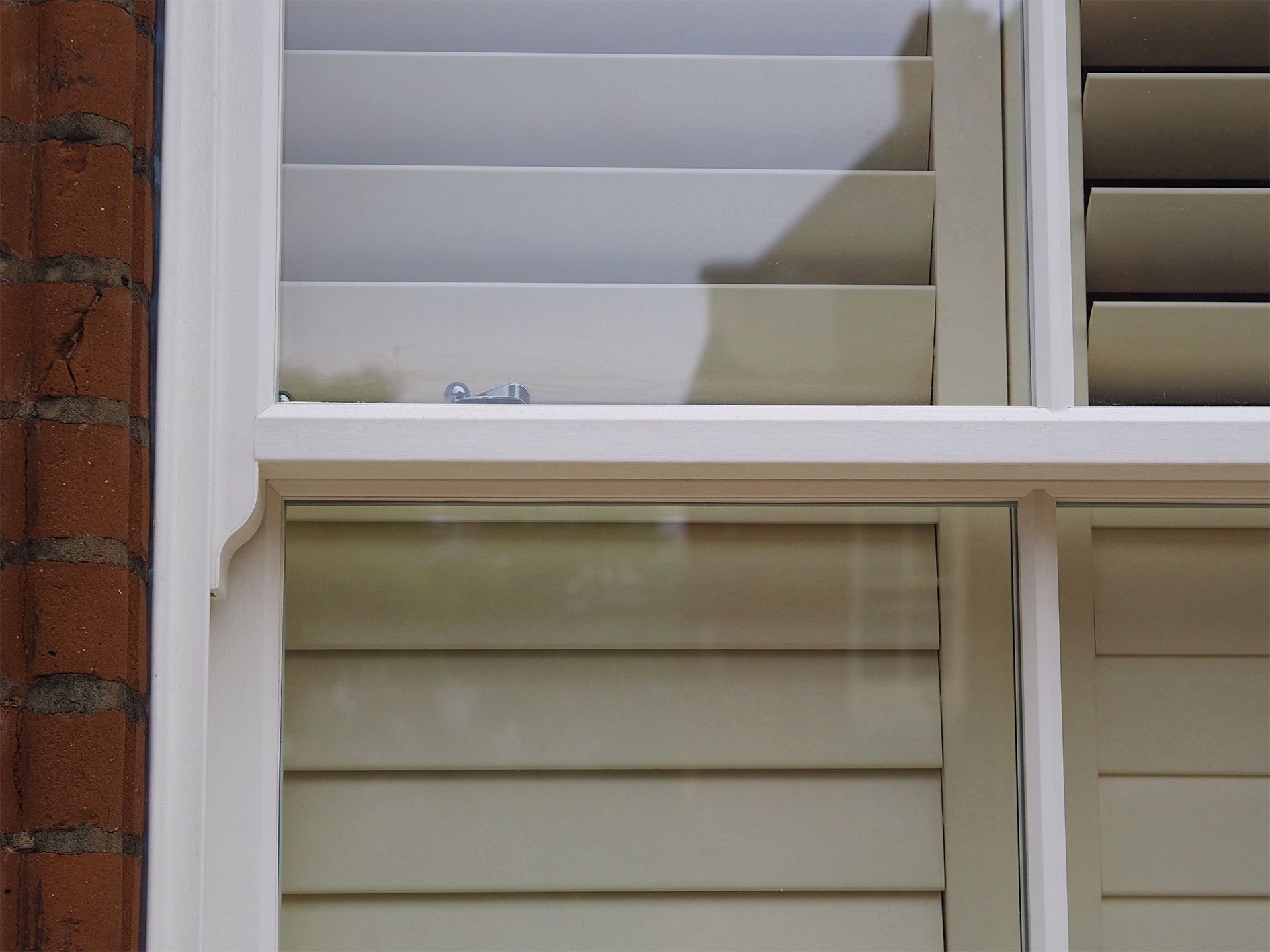 White shutters with angled slats inside a window frame, against a brick wall.