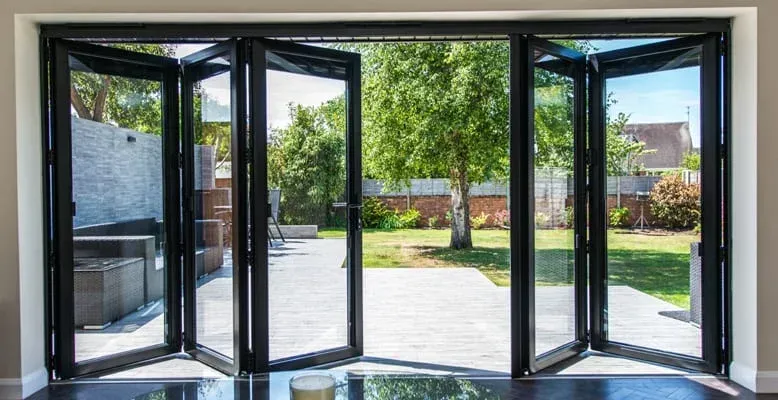 Open folding glass doors lead to a backyard with trees and patio. Black frames.