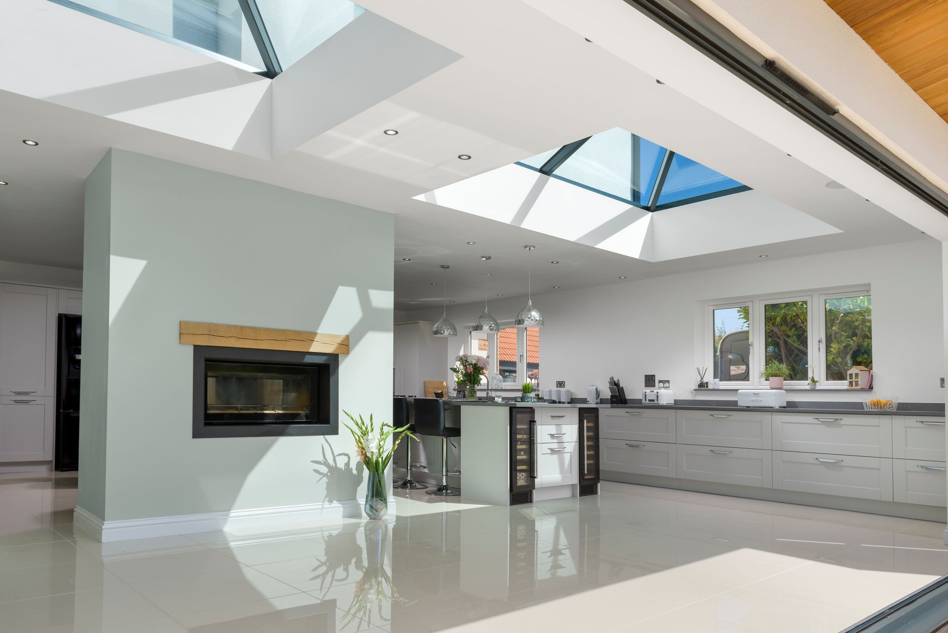Modern kitchen interior with fireplace, skylight, and large sliding doors. Pale blue walls, white floors.