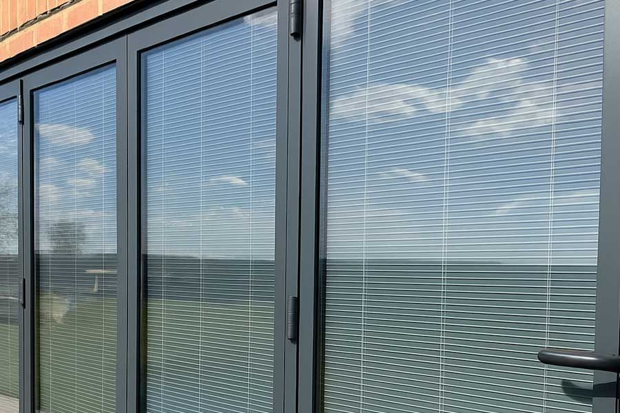 Gray bi-fold doors with blinds reflect a cloudy sky, a brick wall to the left, and a grassy area outside.