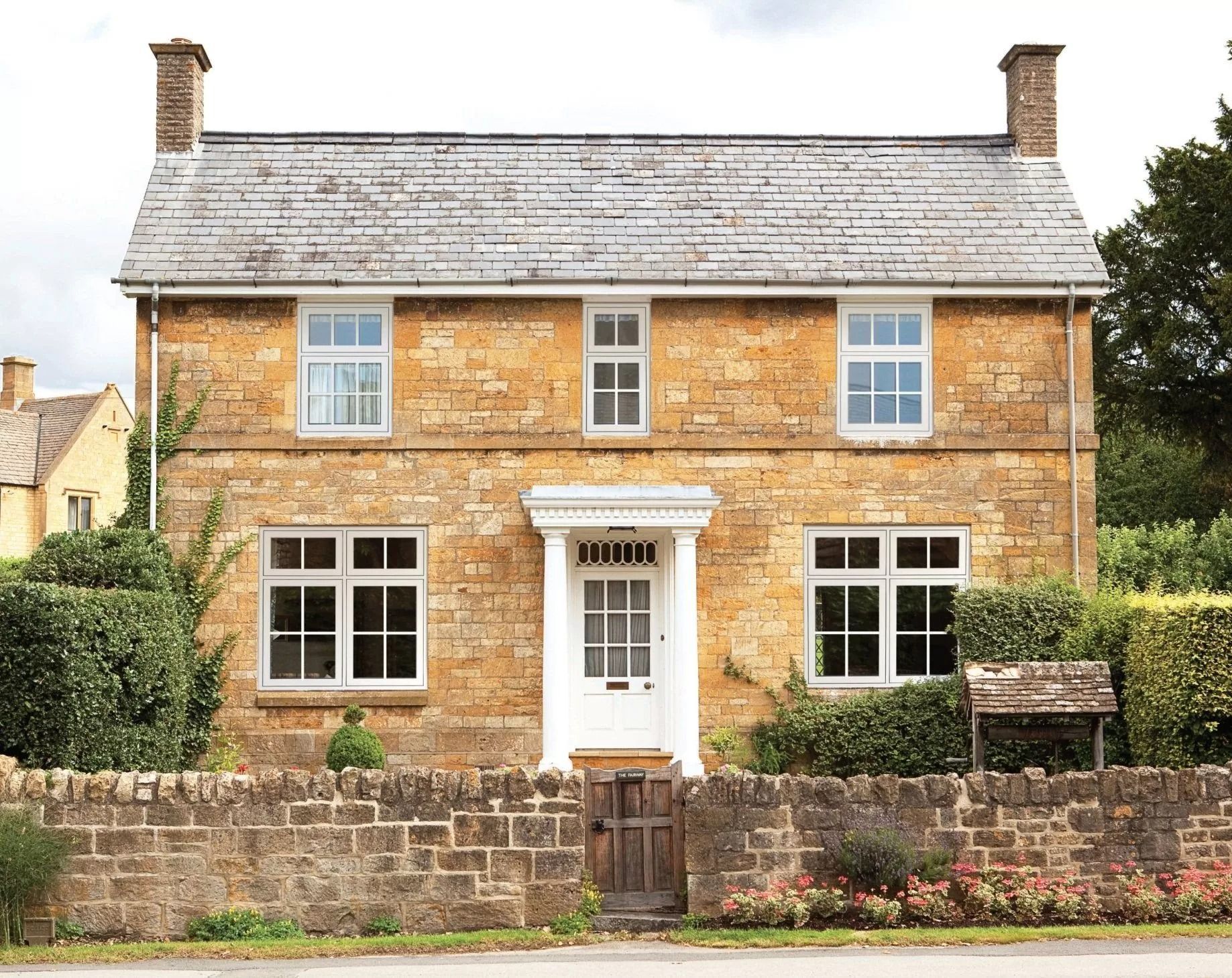 Yellow stone cottage with white windows and door, grey roof, behind a stone wall and hedge.