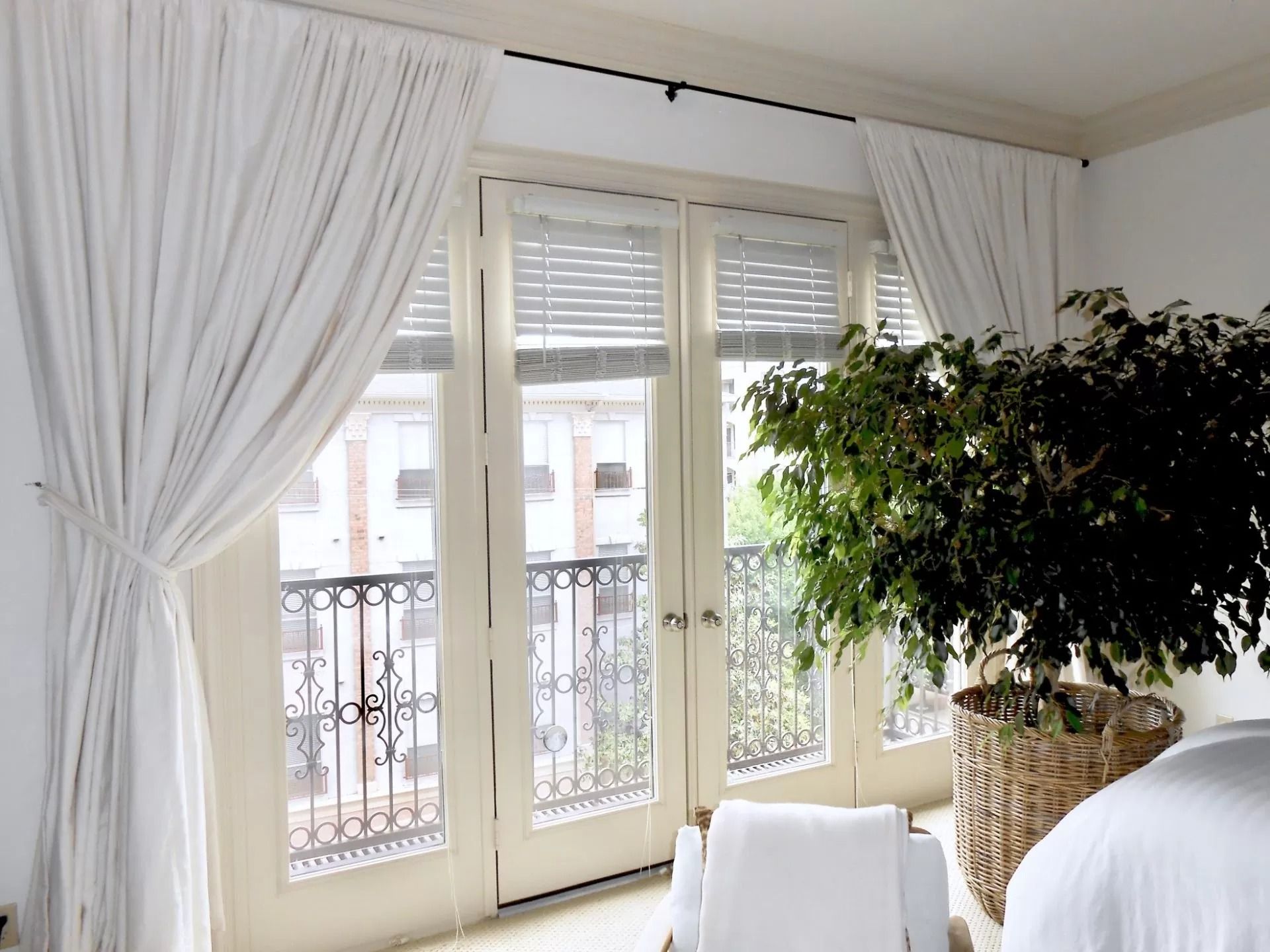 White curtains framing French doors, balcony view, large potted plant, white chair.