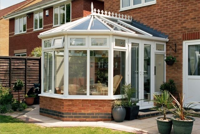 White and glass conservatory with a brick base attached to a house with a red brick exterior.