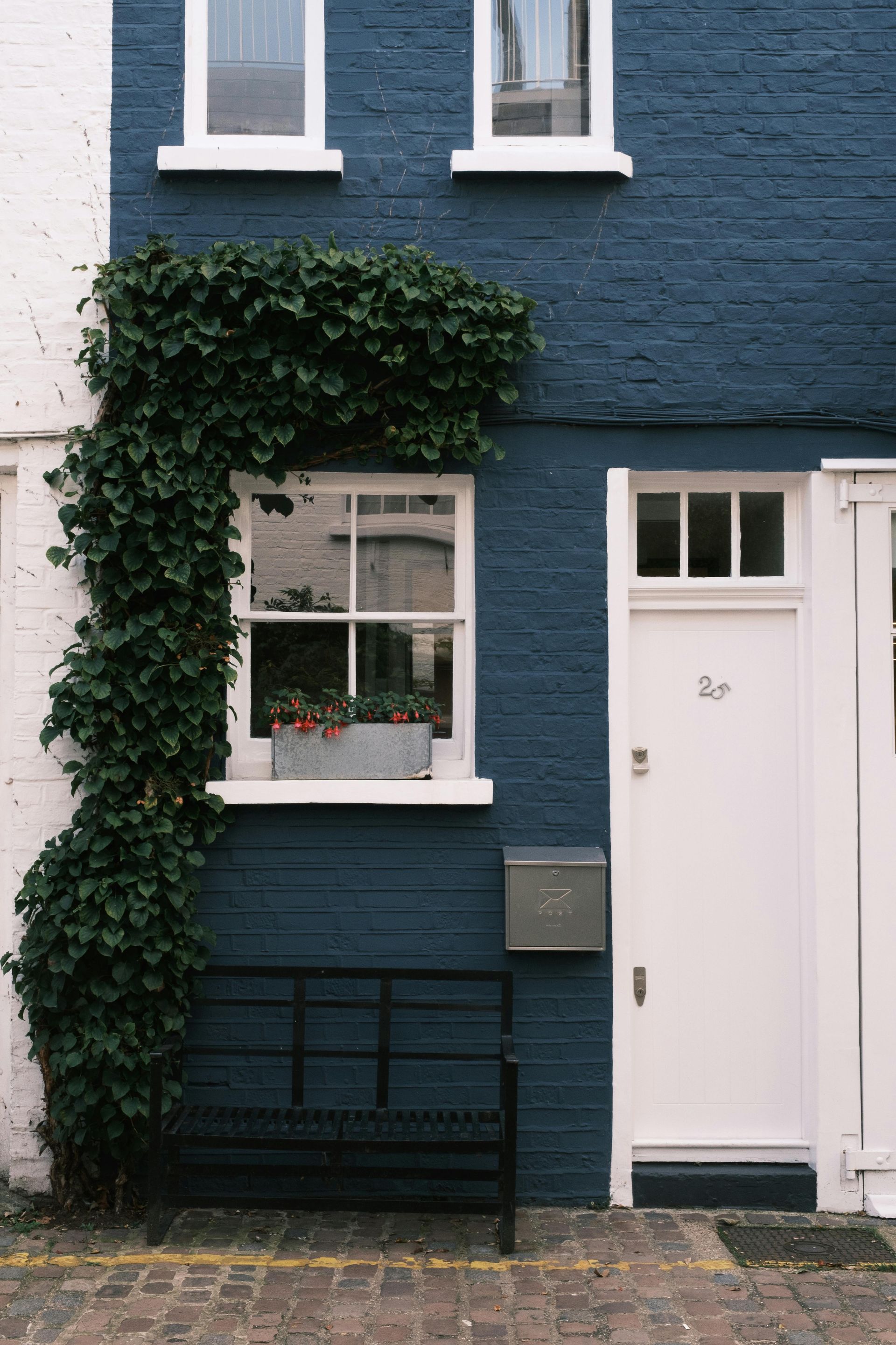 Blue brick house with ivy, white trim, door, window box, bench, and mailbox on cobblestone street.