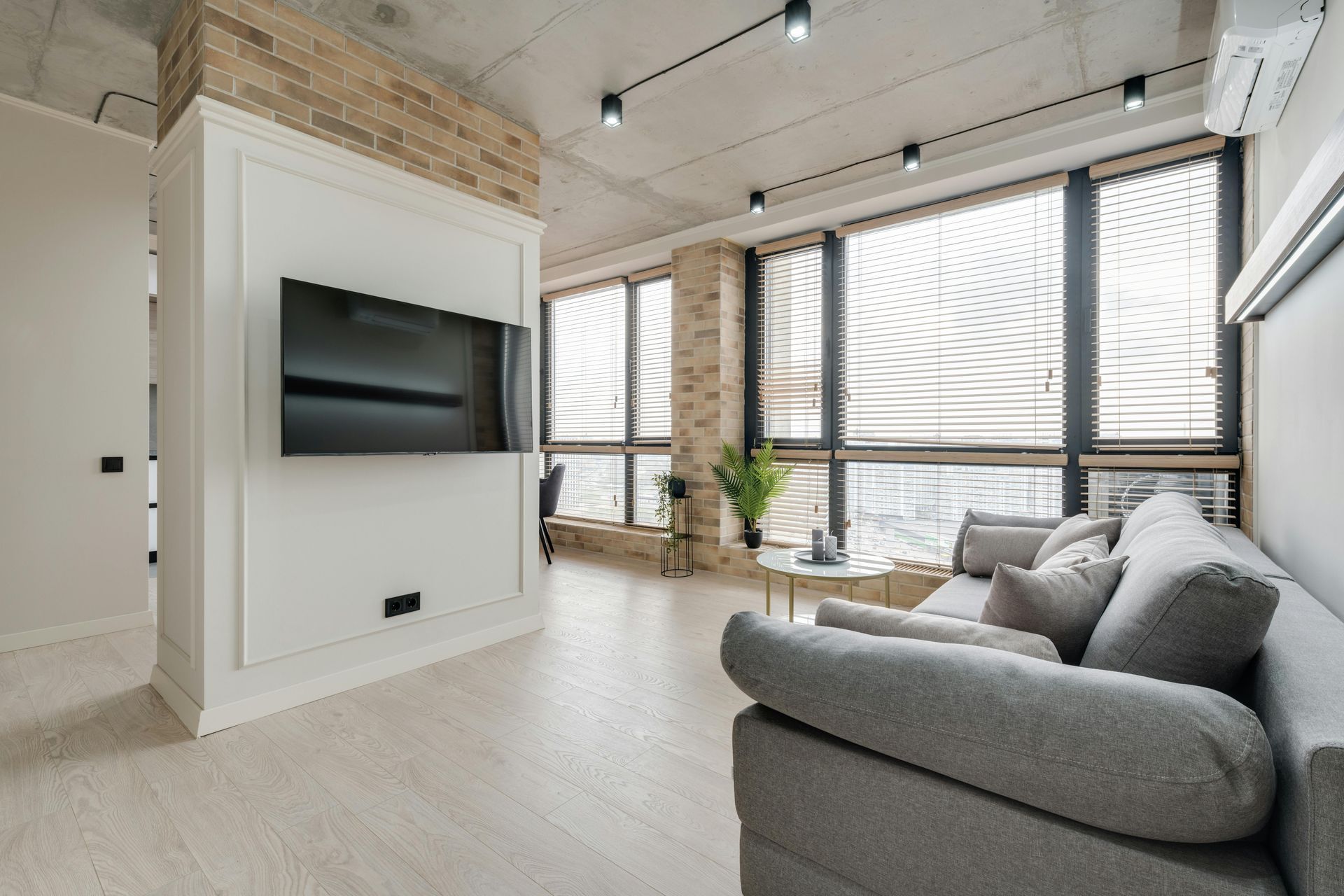 Modern living room with a grey sofa, TV, large windows, and exposed brick.