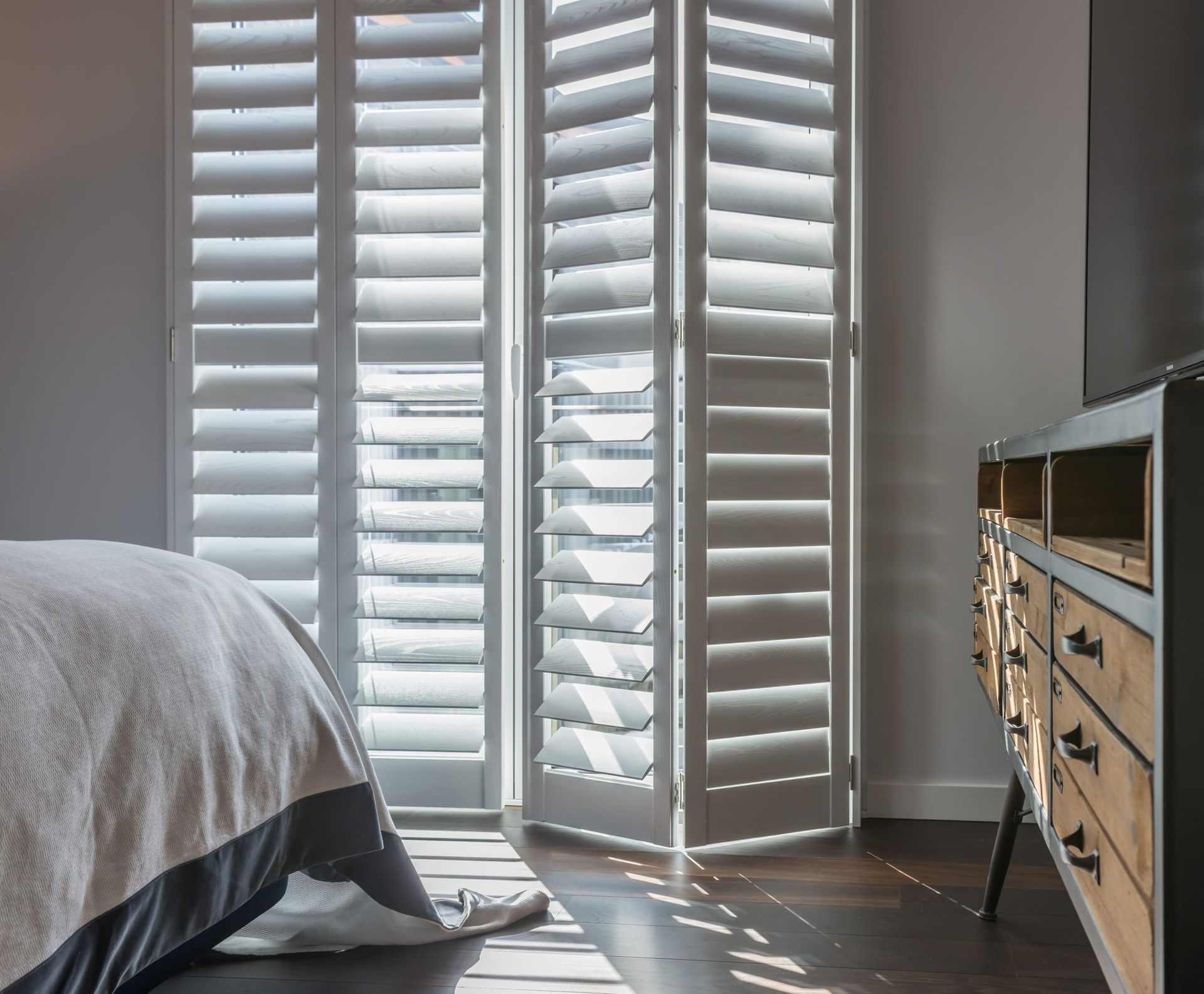Bedroom with white shutters, sunlight, dark wood floor, bed, and wooden cabinet.