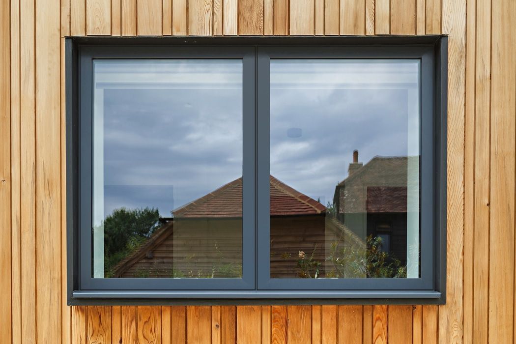 Gray window set in wooden siding, reflecting a cloudy sky and buildings.