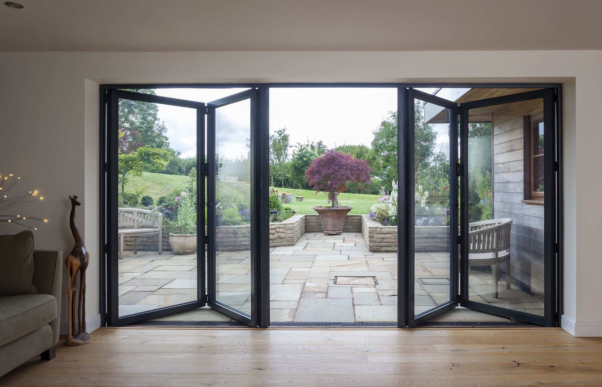 Folding glass doors open onto a stone patio with a garden and tree.