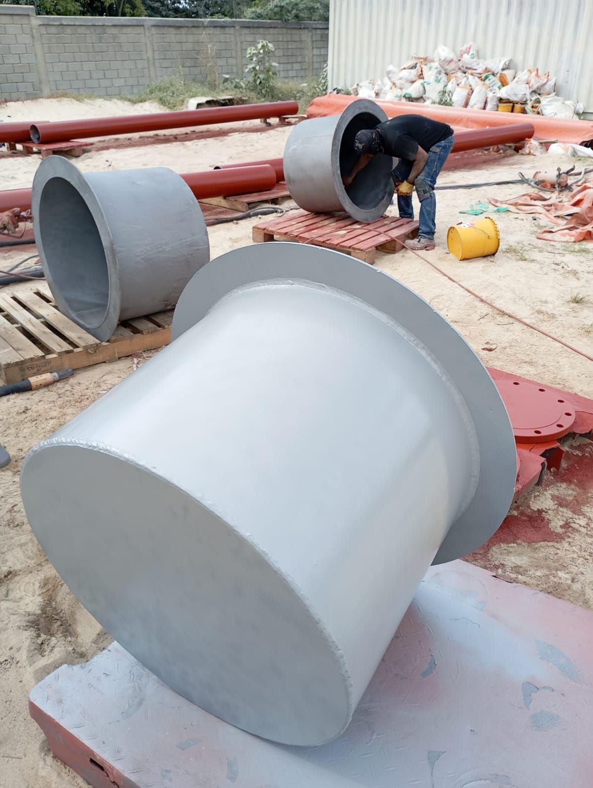 Obrero pintando grandes cilindros metálicos grises en una obra de construcción al aire libre.