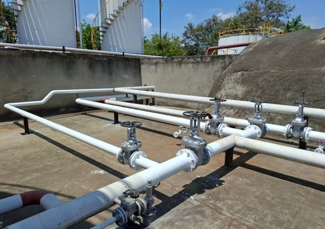 Tuberías y válvulas industriales blancas sobre una azotea de hormigón, junto a grandes depósitos de almacenamiento.