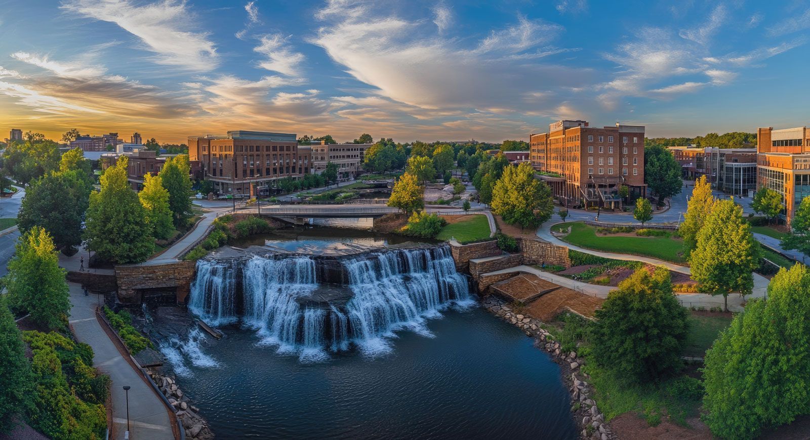Waterfall in a park setting, with buildings and trees. Blue water, sunset sky.