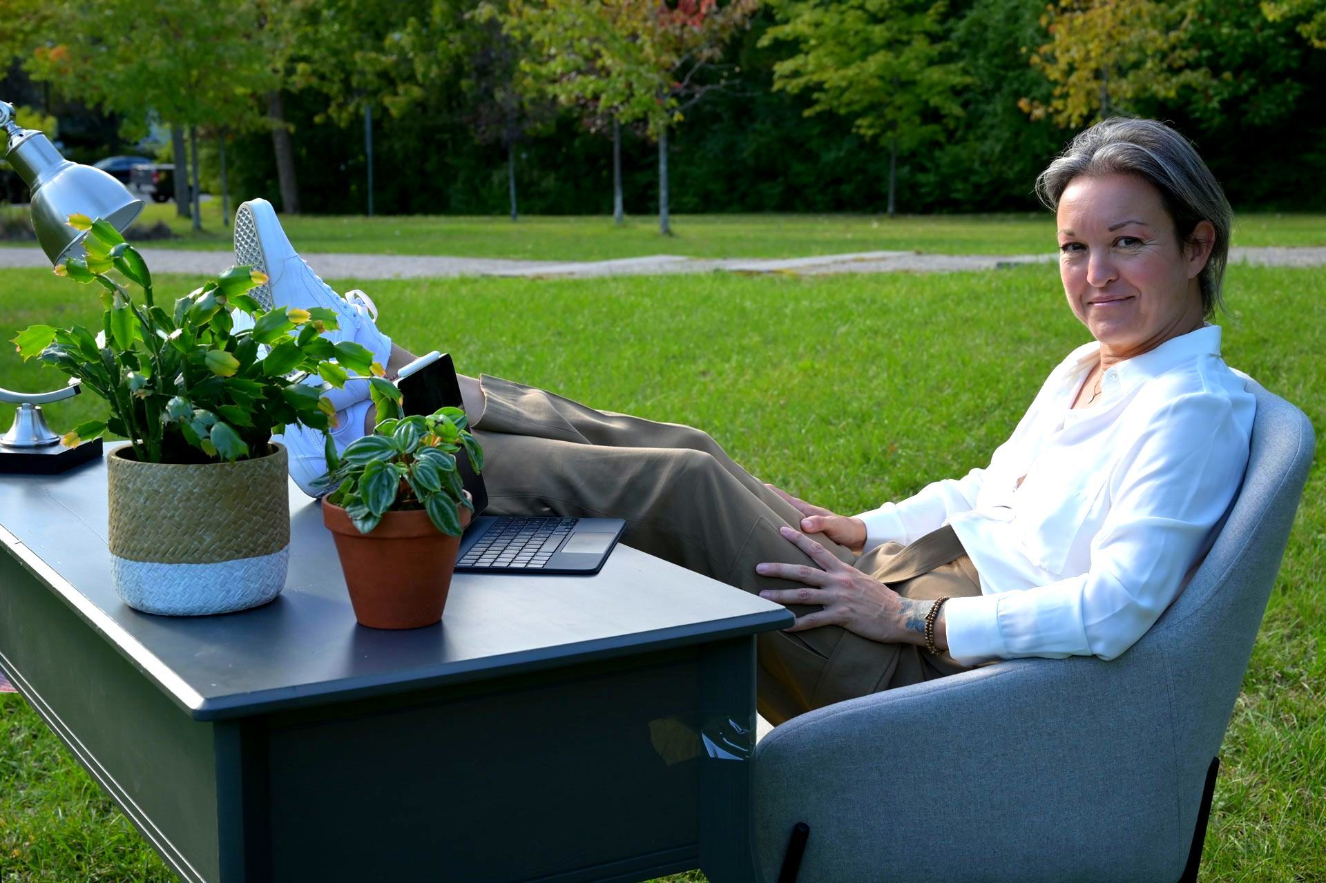 Une femme est assise à un bureau dans l'herbe, les pieds relevés.