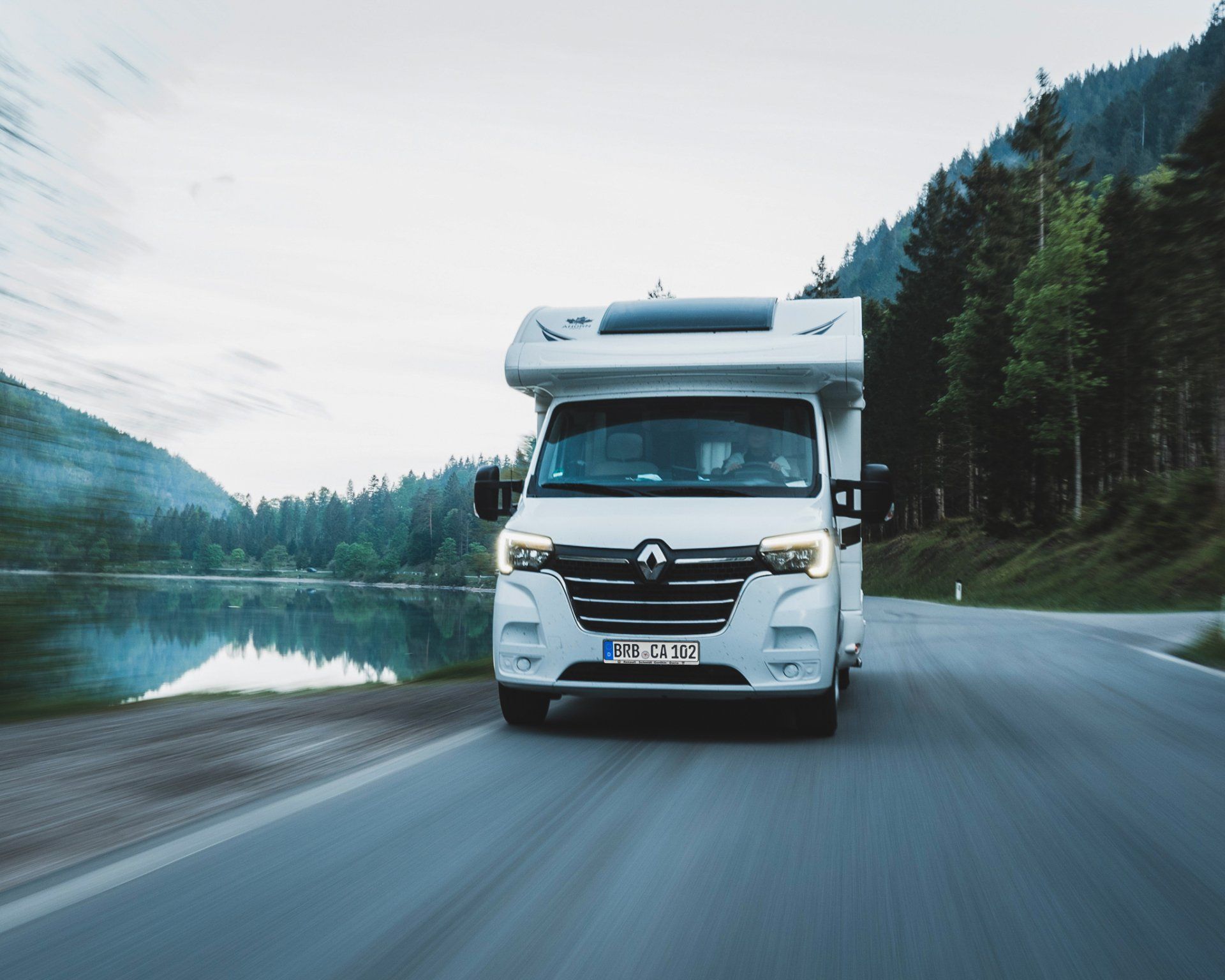 Das Bild zeigt ein weißes Wohnmobil der Marke Renault fährt auf einer kurvigen Landstraße entlang eines ruhigen Sees. Die Scheinwerfer sind eingeschaltet, und das Fahrzeug ist von bewaldeten Bergen umgeben. Die Abend- oder Morgendämmerung taucht die Szene in weiches Licht.