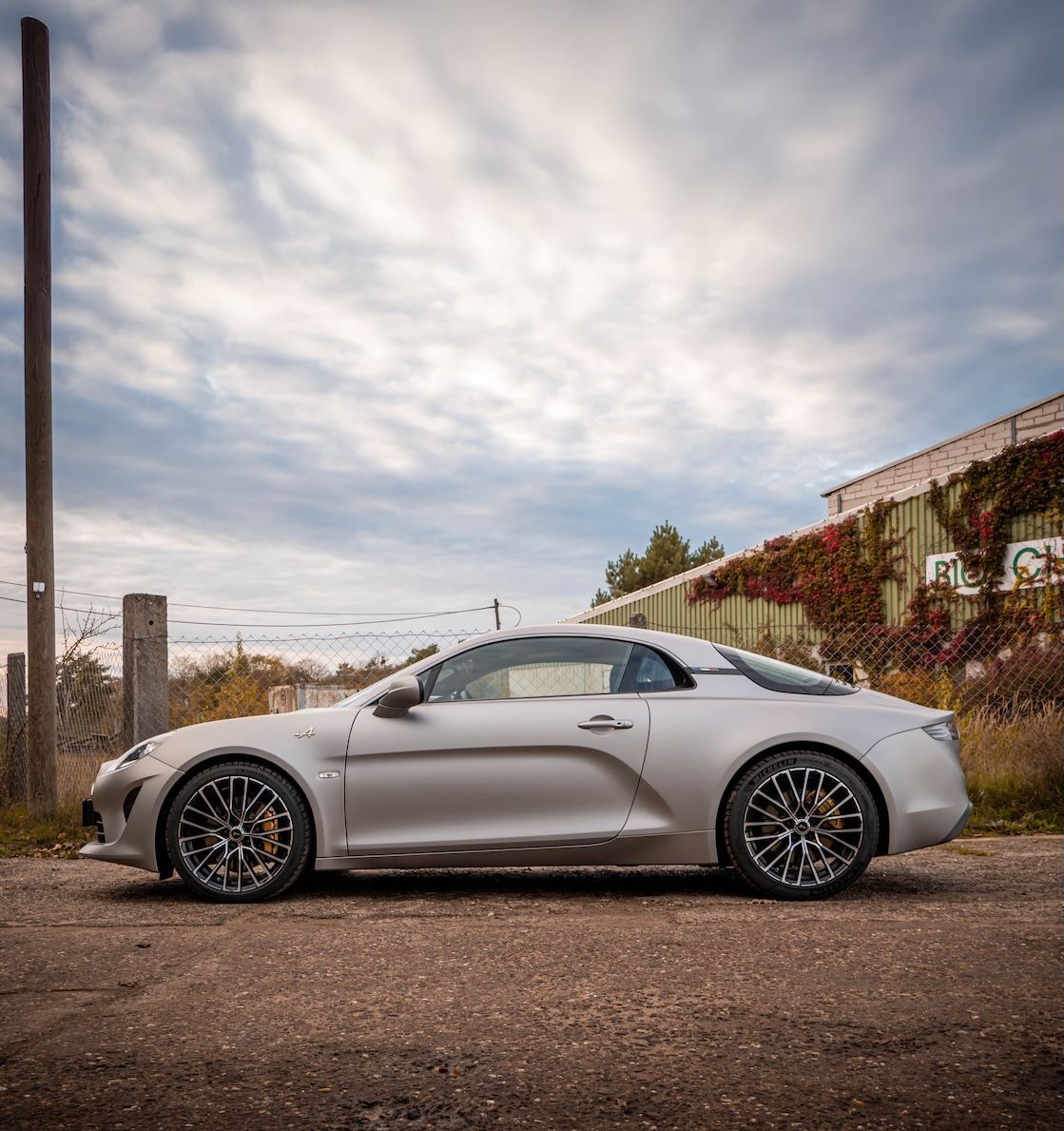 Ein silbernes Sportcoupé der Marke Alpine steht auf einer unbefestigten Straße. Das Fahrzeug hat eine elegante, aerodynamische Form, schwarze Alufelgen mit gelben Bremssätteln und ein geschlossenes Panoramadach. Im Hintergrund sind ein verwittertes Industriegebäude mit grünen Wänden, Rankpflanzen und ein Maschendrahtzaun zu sehen. Der Himmel ist teils bewölkt.