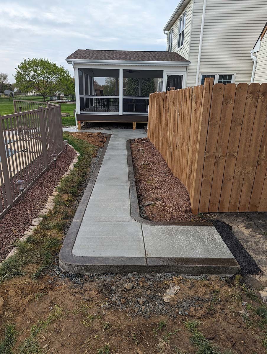 A concrete walkway leading to a screened in porch next to a wooden fence.