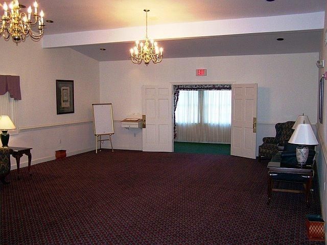 Empty room with burgundy carpet, white walls, and two chandeliers. Exit sign above doorway.