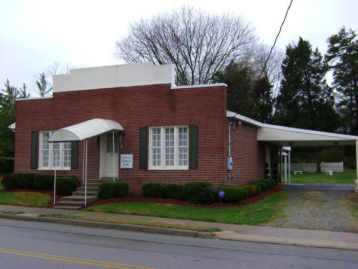 Red brick building with awning over the door and carport on the right. Green grass, shrubbery, and a tree in the background.