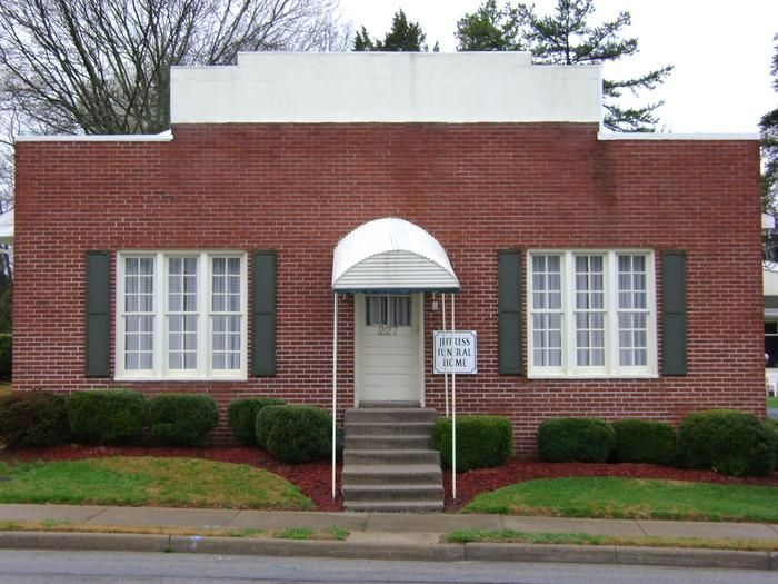 Brick building with white trim, door, awning, two windows, green shutters, shrubs, and sign.