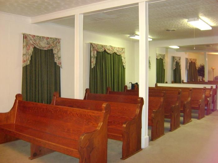 Wooden pews in a room with green curtains and mirrors, possibly a chapel or funeral home.