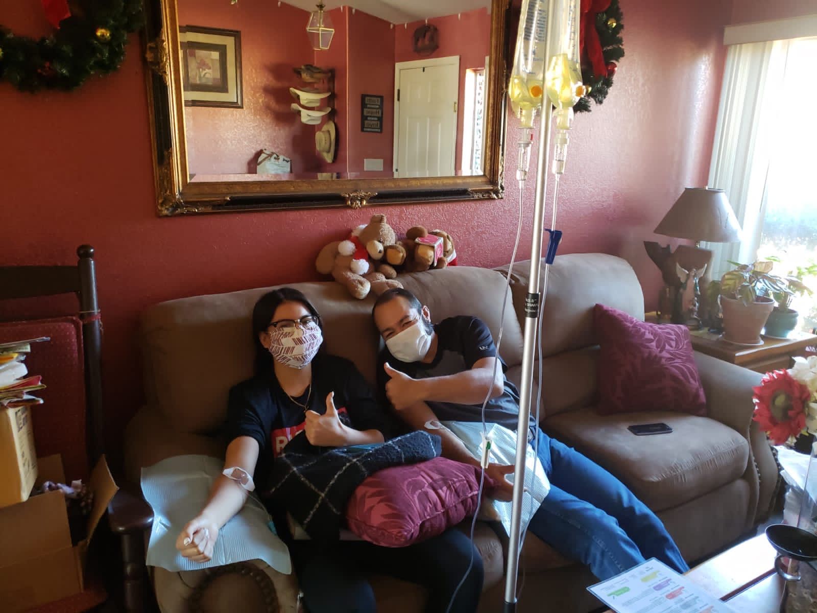 Two people on a couch with IV drips, wearing masks and giving thumbs up in a living room.