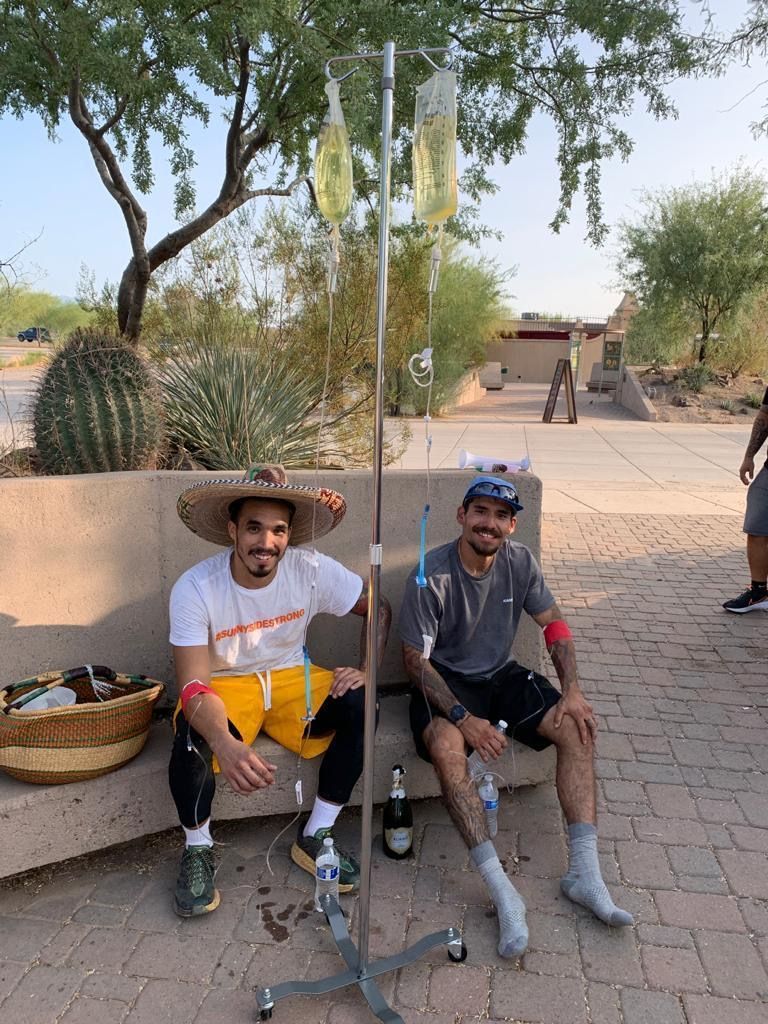 Two men are sitting on a bench with iv bags in their legs.