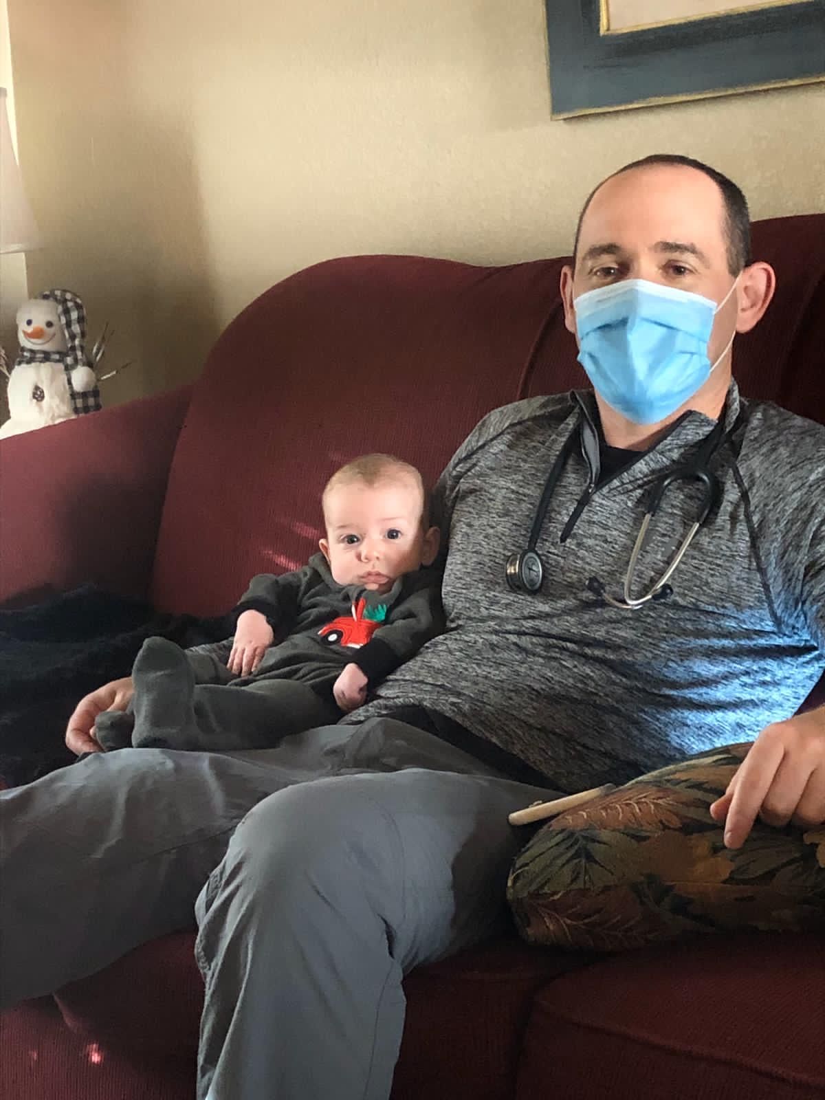 A man wearing a mask is sitting on a couch holding a baby.