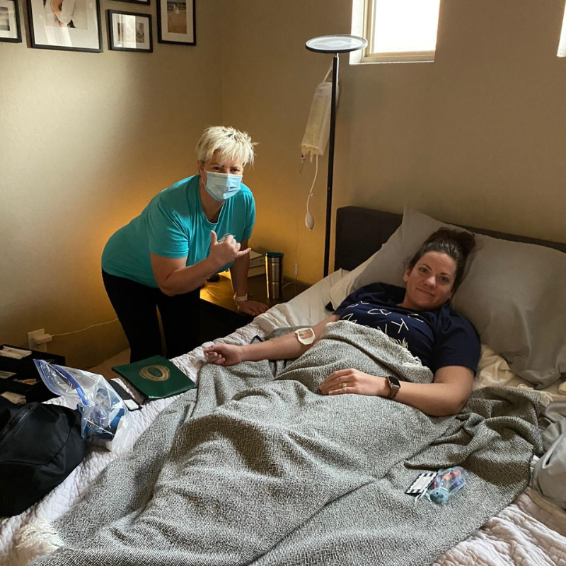 A woman wearing a mask is standing next to a woman laying on a bed