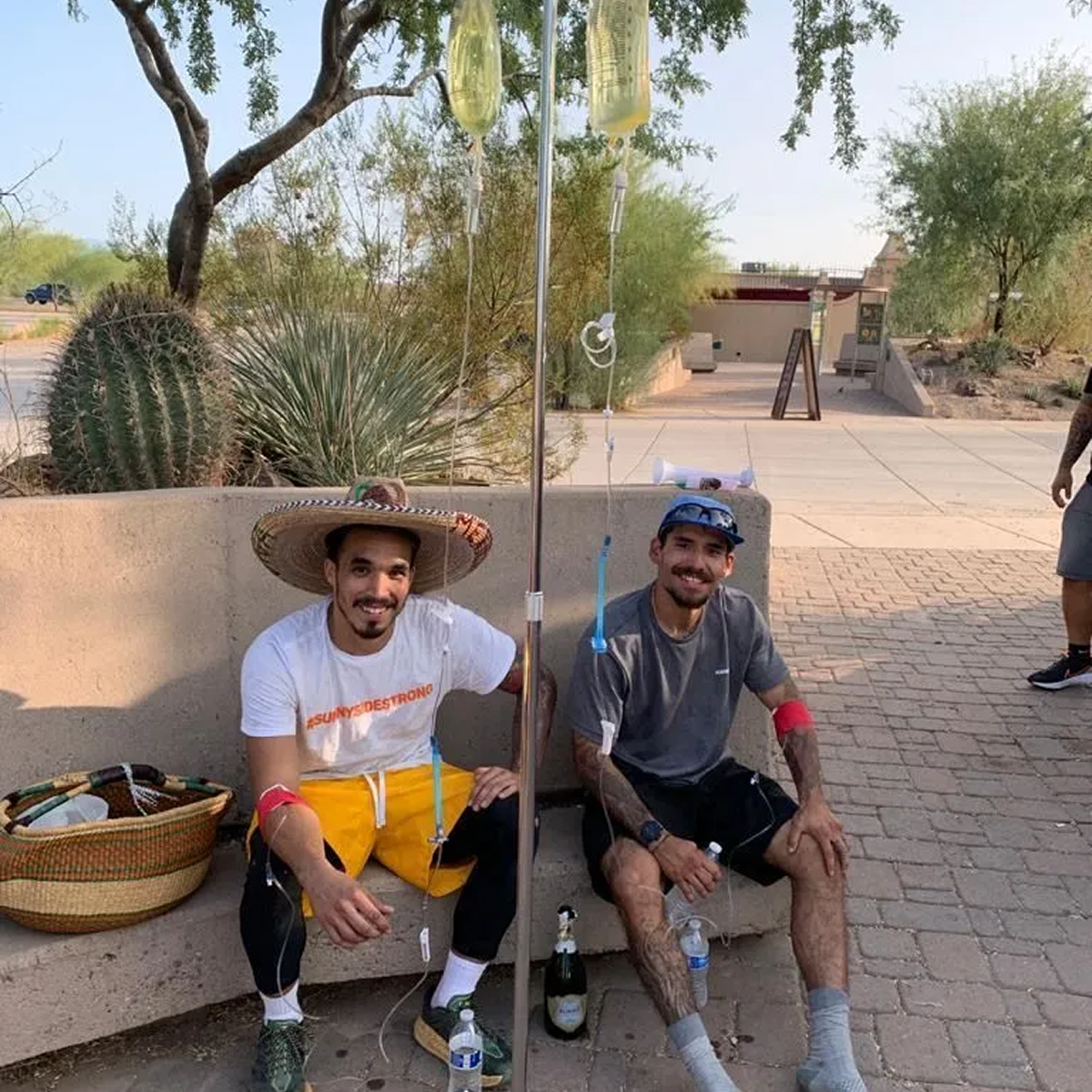 Two men are sitting on a bench with iv bags in their legs.