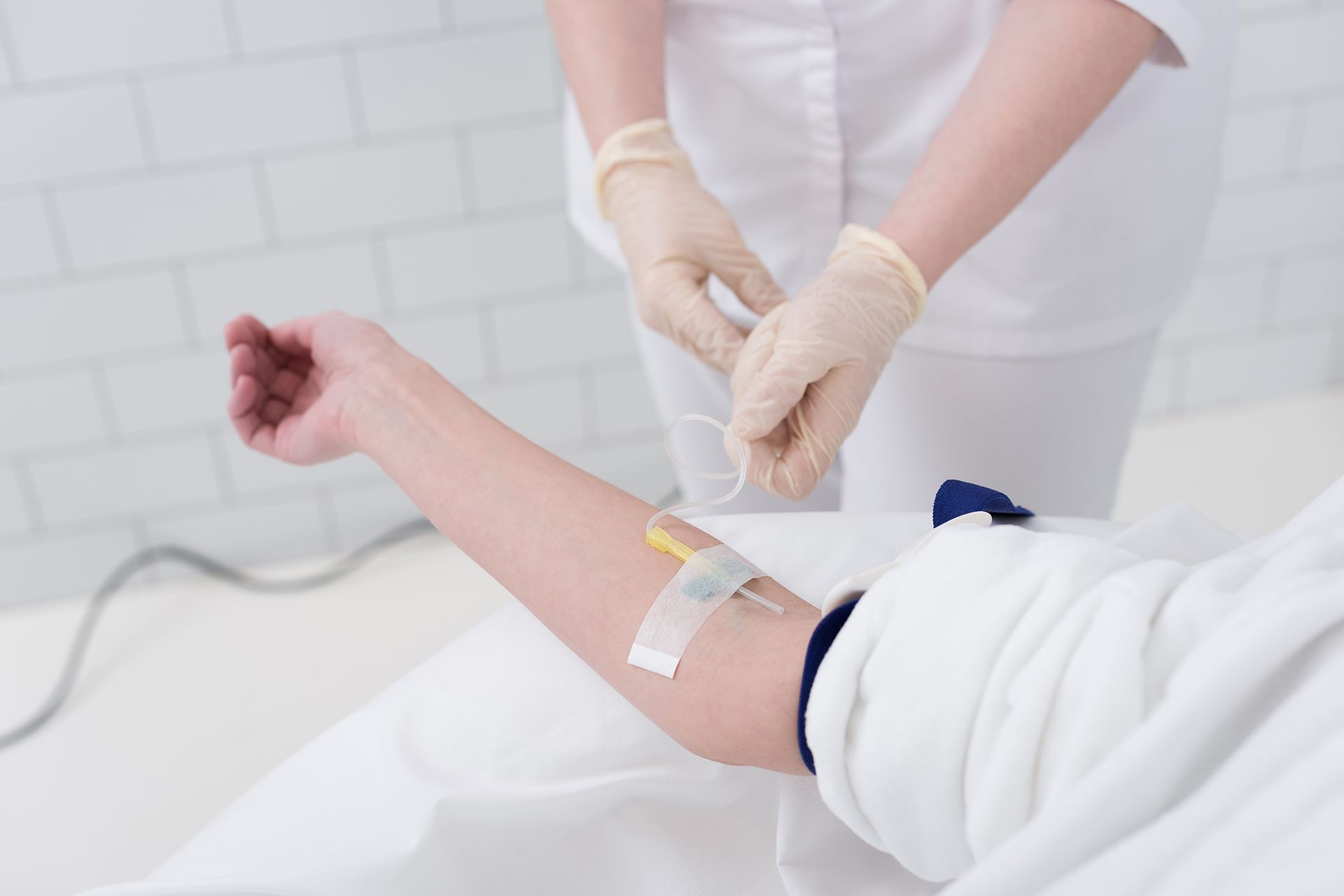 A medical professional in white attire and gloves prepares an IV line on a patient's arm in a clinical setting.