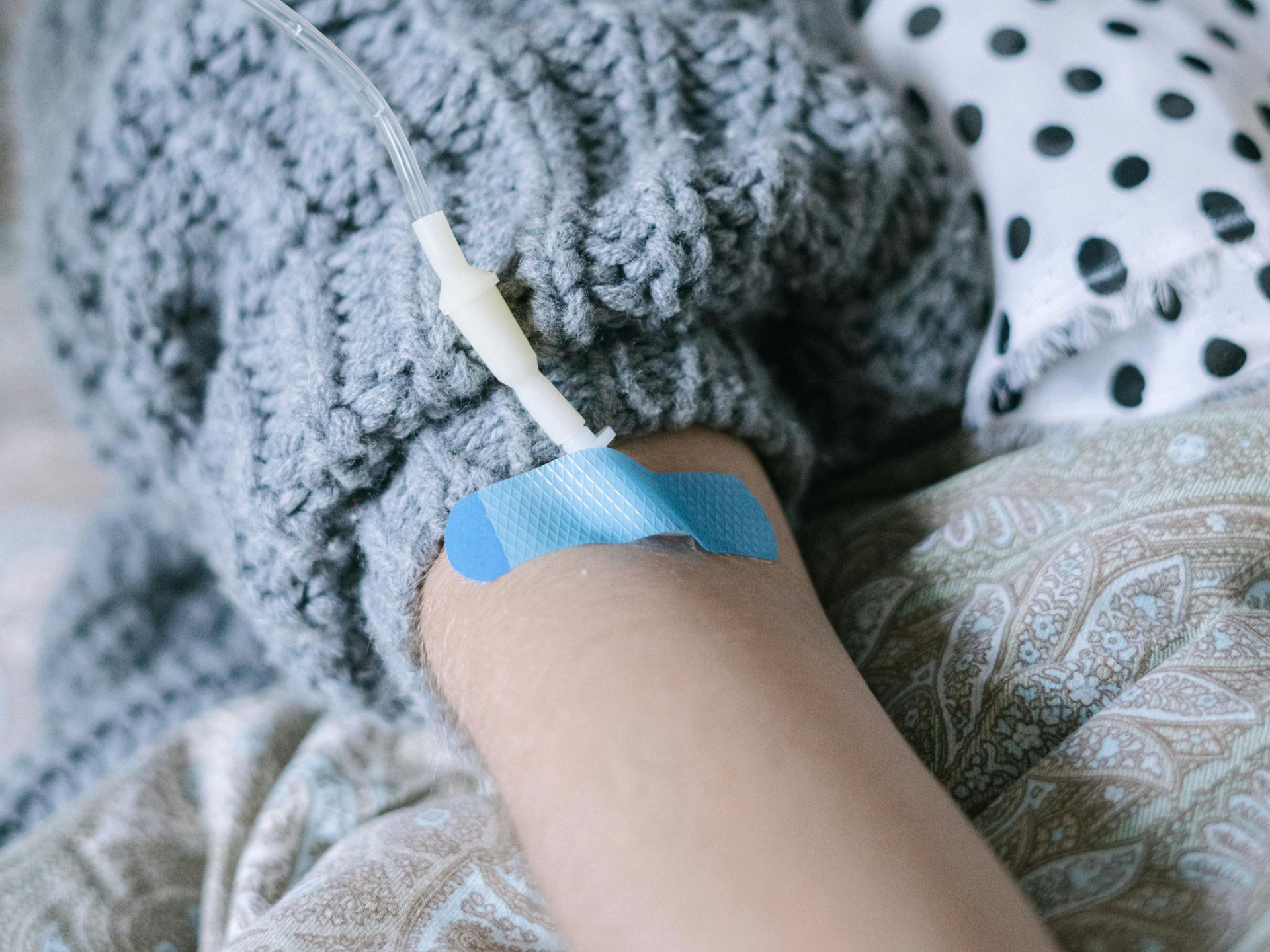 A person's arm with an IV line secured by blue medical tape, resting on bedding while wearing a gray knit sweater.