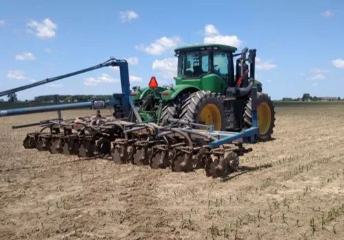 Green tractor pulling a large farm implement across a dry field under a blue sky