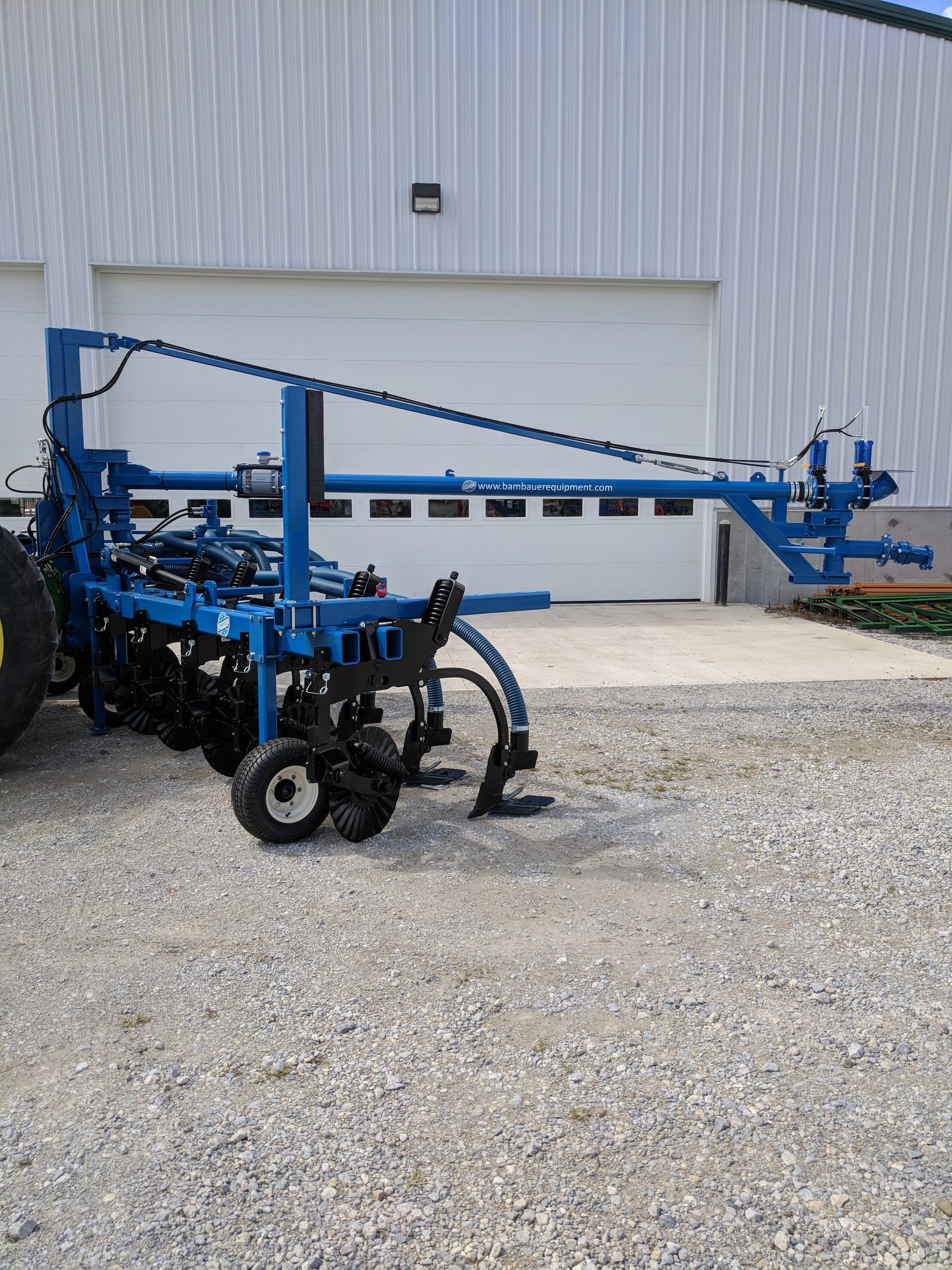 Blue agricultural sprayer attached to a tractor parked on gravel beside a white metal building.