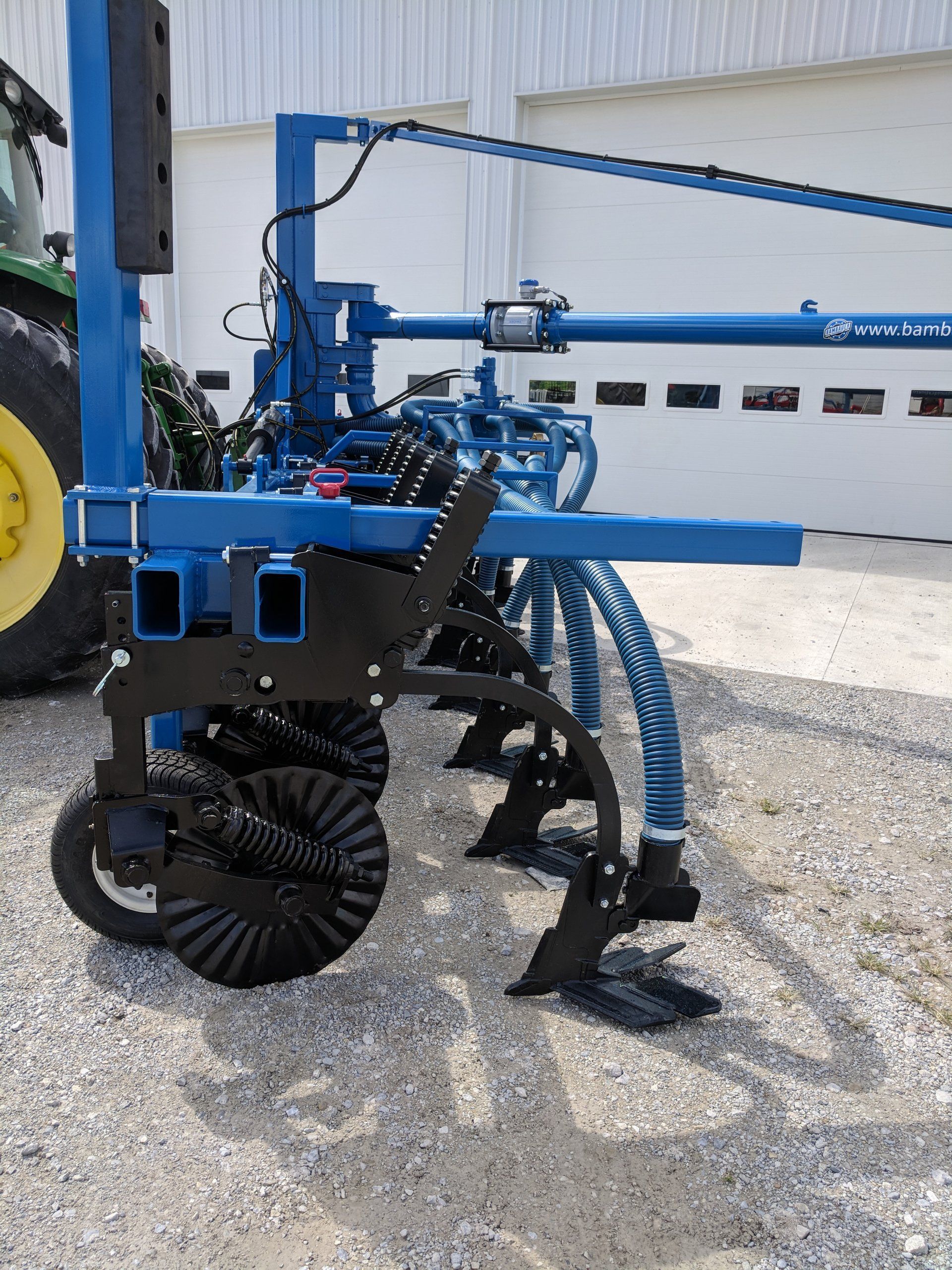 Blue agricultural machine with spiked wheel attachment parked on gravel beside a building.