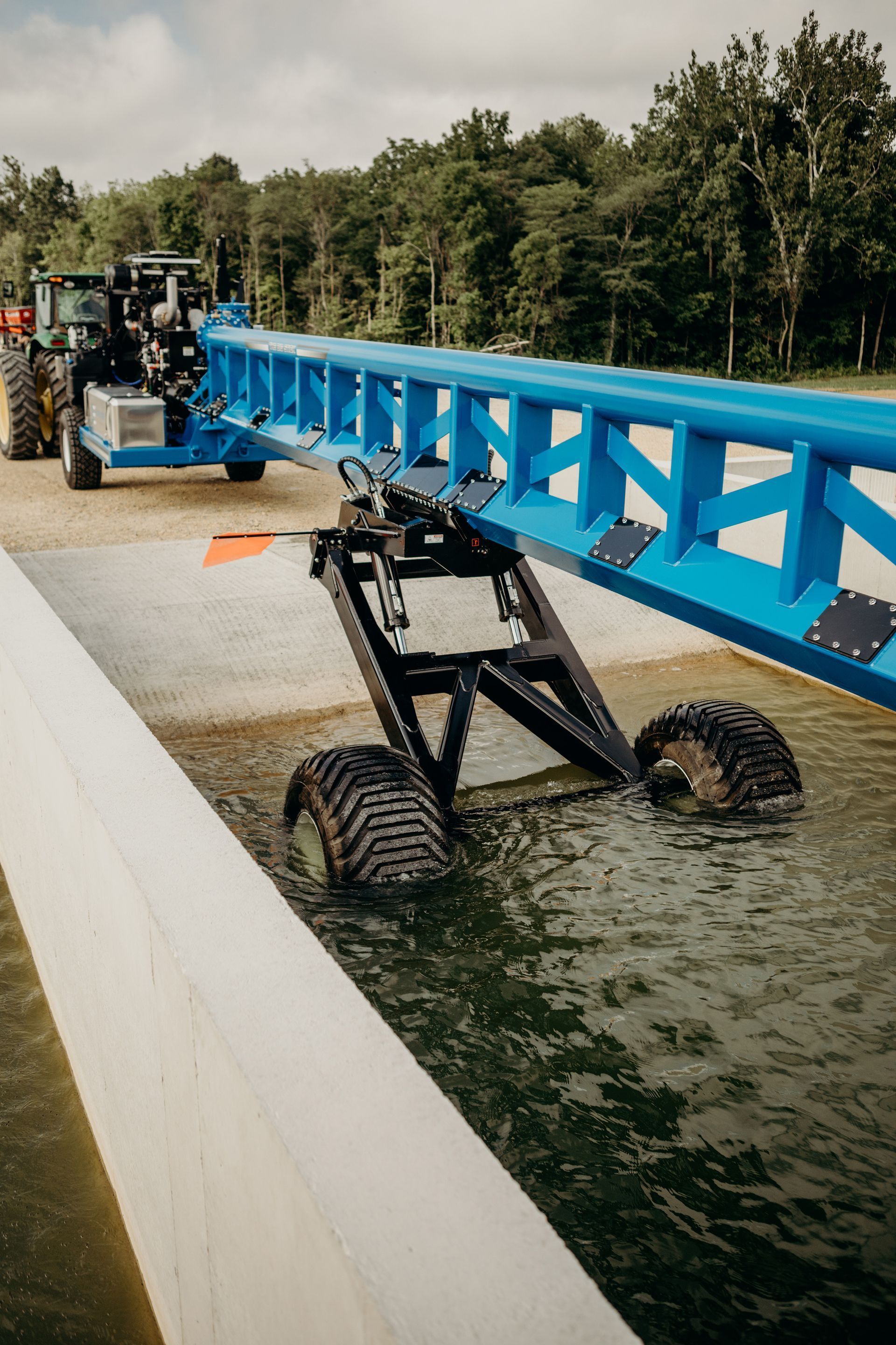 Blue bridge-launching machine on water beside a canal, with tractor and trees in the background
